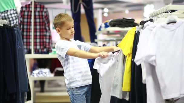 Милый улыбающийся мальчик стоит рядом с одеждой и выбирает — стоковое видео