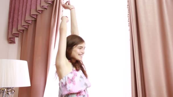 Young joyful woman holding the curtains open to look out of large light window at home, turning to look and smile at camera, interior. Aspirational lifestyle indoors. negligee — Stock Video