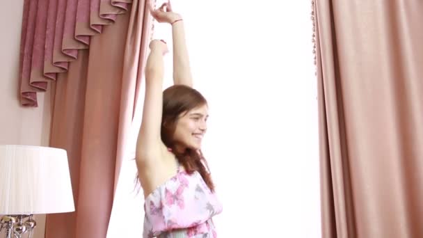 Jovem mulher alegre segurando as cortinas abertas para olhar para fora da grande janela de luz em casa, transformando-se para olhar e sorrir para a câmera, interior. Estilo de vida inspirador dentro de casa. insignificante — Vídeo de Stock