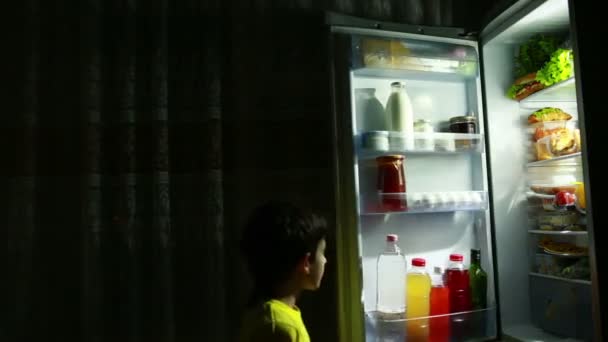 Niño comiendo un bocadillo frente al refrigerador en medio de la noche. niño hambriento — Vídeo de stock