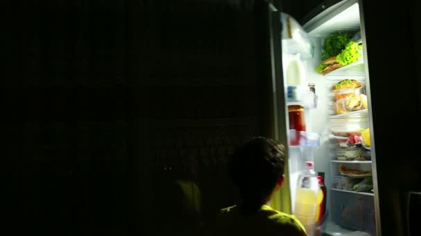 Niño comiendo un bocadillo frente al refrigerador en medio de la noche. Chico hambriento. leche para beber — Vídeo de stock