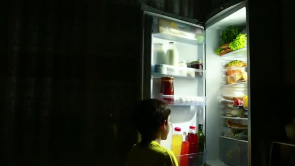 Niño comiendo un bocadillo frente al refrigerador en medio de la noche. Chico hambriento. alas de pollo — Vídeo de stock