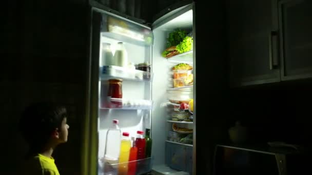 Criança comendo um lanche na frente da geladeira no meio da noite . — Vídeo de Stock