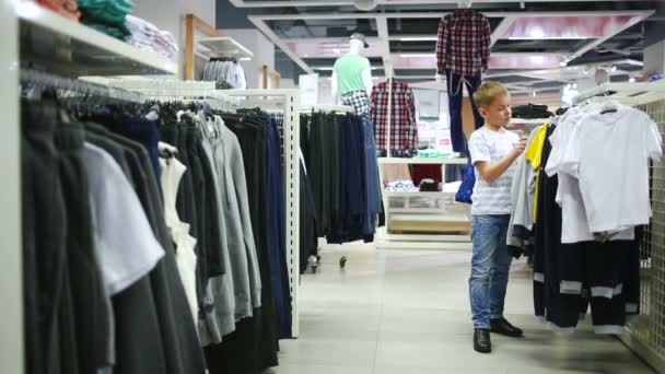 Bonito menino sorridente fica perto de roupas e escolher — Vídeo de Stock