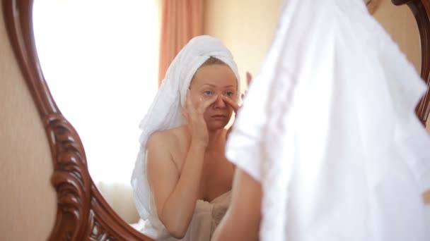 Mujer aplicando mascarilla hidratante crema para la piel en la cara mirando en el espejo . — Vídeo de stock