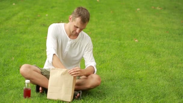 Guy having lunch on meadow. man eating a sandwich on the lawn and drinks juice. — Stock Video