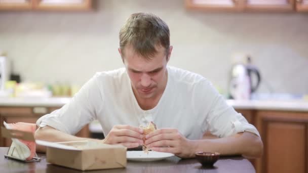 Όμορφος άνδρας μάσημα και απολαμβάνοντας ένα χάμπουργκερ με πατάτες στο σπίτι — Αρχείο Βίντεο