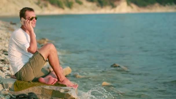 Hombre sonriente sentado en la playa y hablando en un teléfono inteligente . — Vídeos de Stock