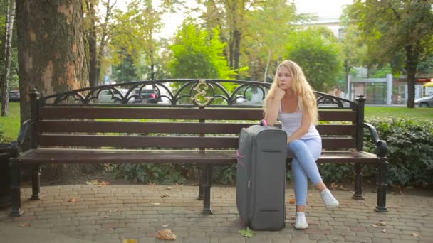 Ledsen flicka sitter på bänken med en resväska. — Stockvideo