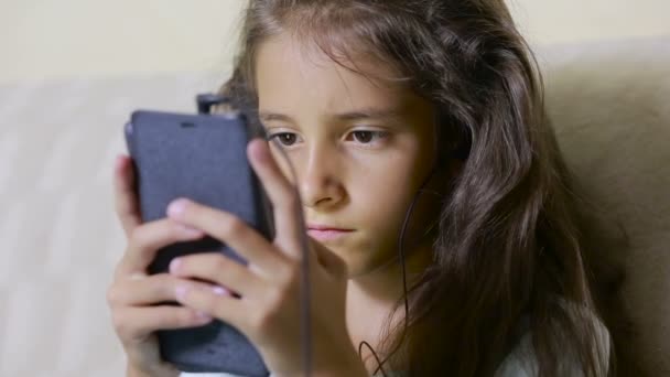 Κορίτσι παιδί με ένα κινητό τηλέφωνο πραγματοποιεί αναζήτηση στο Internet. ακουστικά — Αρχείο Βίντεο