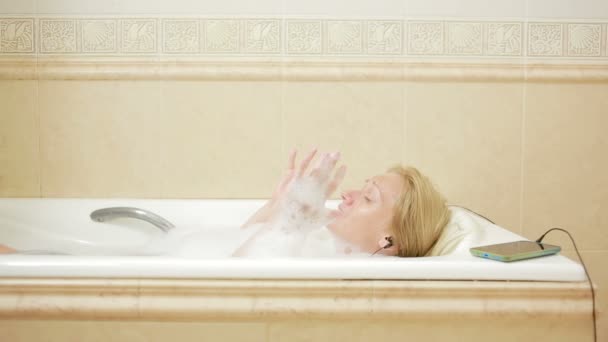 Hermosa mujer tomando un baño y escuchando música en los auriculares. teléfono celular — Vídeo de stock