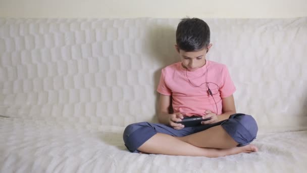 Niño con smartphone y auriculares escuchando música o jugando en casa — Vídeos de Stock