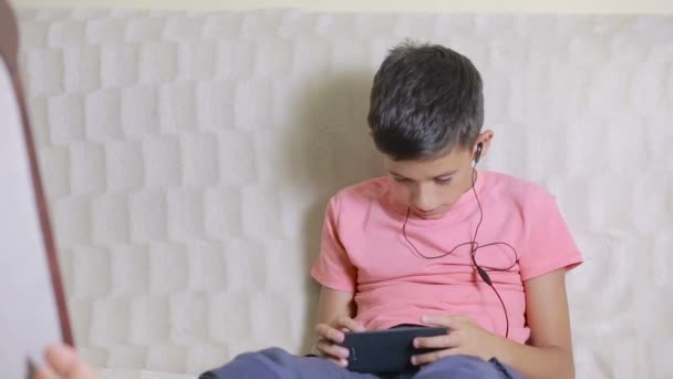 Pojke med smartphone och hörlurar lyssnar på musik eller spelar spel hemma — Stockvideo