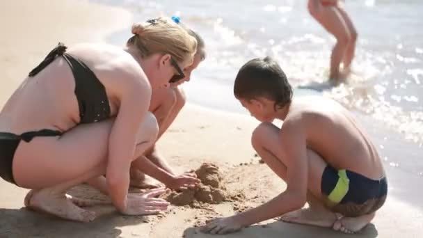 Matka z dziećmi na plaży. zbudować zamek z piasku. chłopiec i dziewczynka — Wideo stockowe