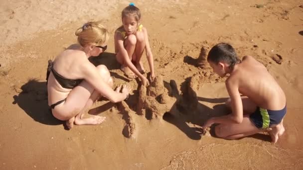 Matka z dziećmi na plaży. zbudować zamek z piasku. chłopiec i dziewczynka — Wideo stockowe