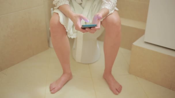 बाथरूममध्ये शौचालयात बसलेली स्त्री. स्मार्टफोन वापरणे. घर बाथरोब — स्टॉक व्हिडिओ