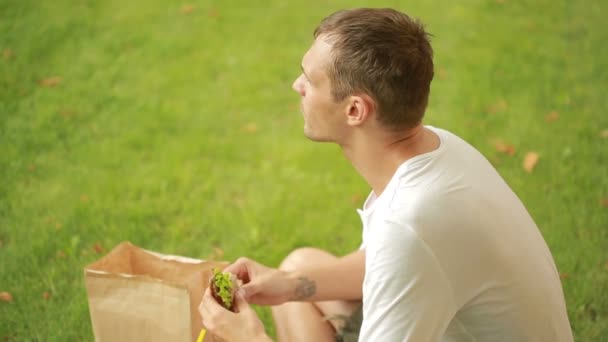 Um tipo a almoçar no prado. homem comendo um sanduíche no gramado e bebe suco . — Vídeo de Stock