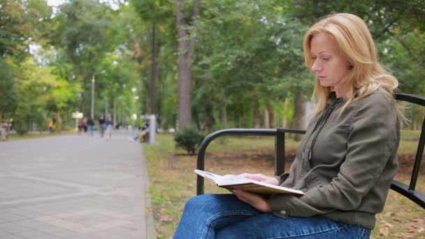 Блондинка жінка читання книги на Brench в Осінній Парк. — стокове відео