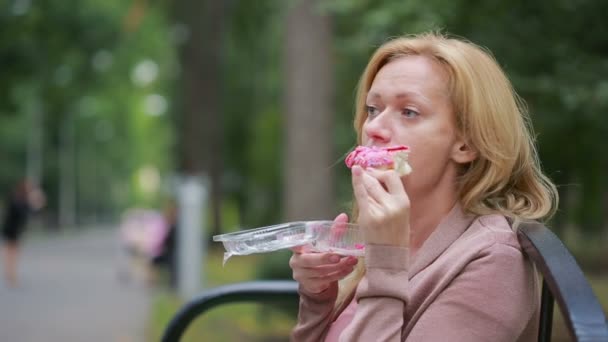 Uma loira num parque a comer um donut. Mulher ao ar livre mastiga pão doce — Vídeo de Stock
