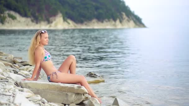 青い透明な海海の水の近くの岩に座ってビキニでセクシーな美女. — ストック動画
