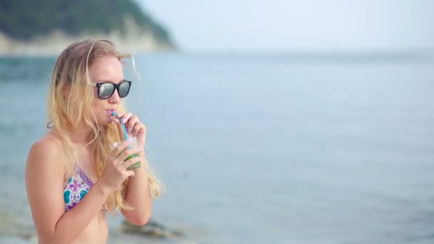 Une fille en bikini boit du jus à travers une paille. femme à la mer . — Video