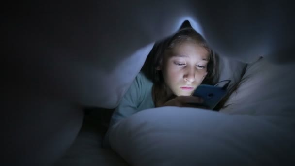 Gadis kecil menggunakan ponsel di malam hari. Bersembunyi di bawah selimut — Stok Video