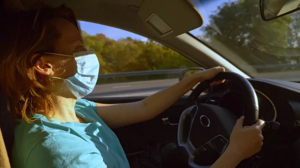 A woman in a t-shirt and a medical mask is driving a car — Stock Photo, Image