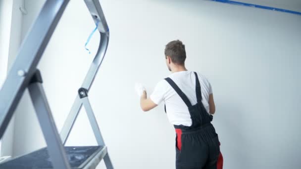 Un hombre en traje de trabajo pinta una pared blanca con pintura blanca con sus manos — Vídeo de stock