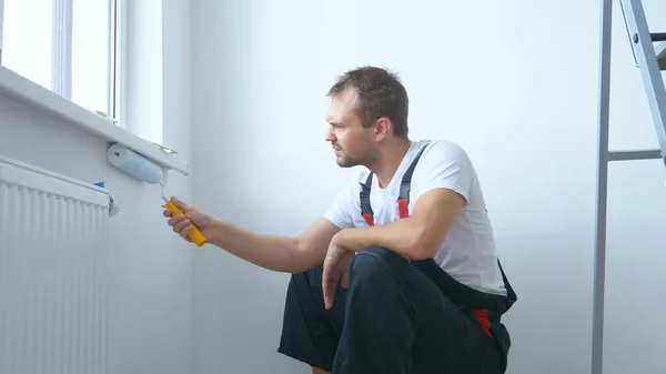 Knappe man in het werk overalls verft de muur met een roller witte verf — Stockfoto