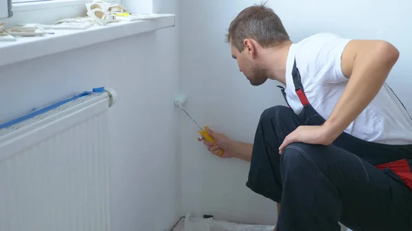 Homem bonito no macacão de trabalho pinta a parede com uma pintura branca rolo — Fotografia de Stock