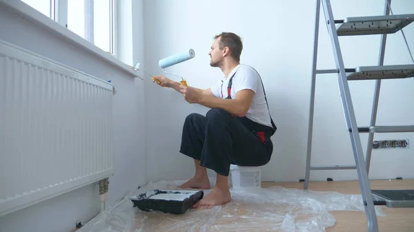 Knappe man in het werk overalls verft de muur met een roller witte verf — Stockfoto