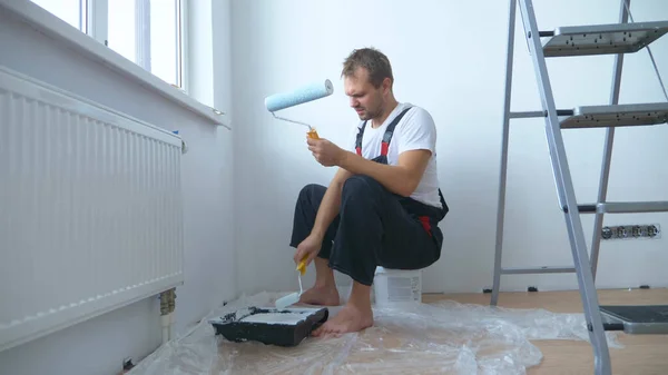 Homem bonito no macacão de trabalho pinta a parede com uma pintura branca rolo — Fotografia de Stock