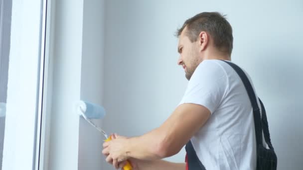 Hombre guapo en overoles de trabajo pinta la pared con una pintura blanca del rodillo — Vídeo de stock