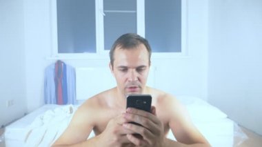 Çıplak bir adam boş bir odada yatağında telefonunu kullanıyor.
