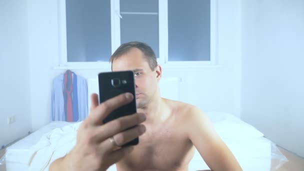 A naked man lies on a mattress in an empty room, using his phone — Stock Video