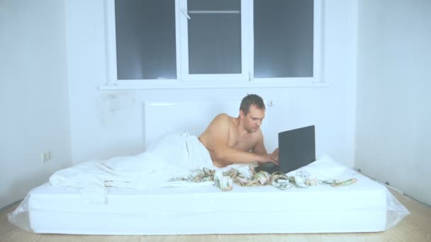 ノートパソコンを使って空き部屋にお金のあるマットレスの上に裸の胴を持つ男 — ストック動画