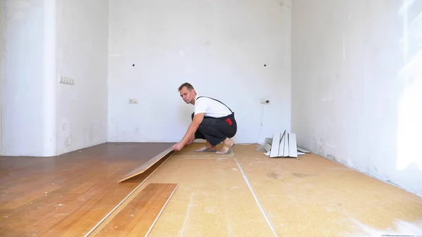 Seorang pria pembangun di overall akan membongkar lantai laminasi tua di sebuah apartemen Stok Foto Bebas Royalti