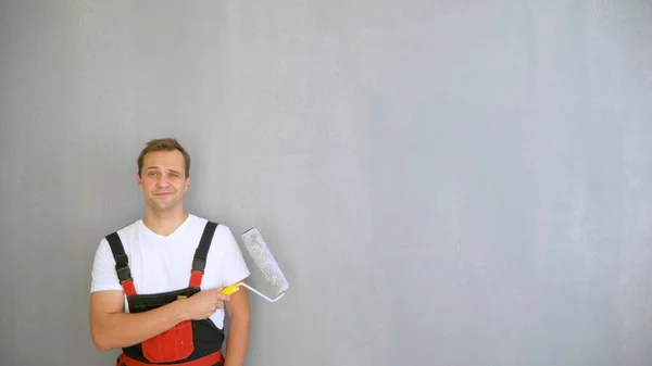 Um homem bonito em macacão de trabalho com um rolo de tinta olhando para a câmera — Fotografia de Stock