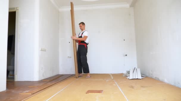 Um homem construtor em macacão vai desmontar um piso laminado velho em um apartamento — Vídeo de Stock