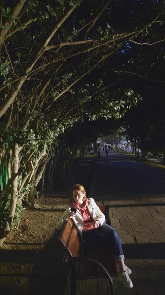 Vrouw in wit lederen jas gebruik smartphone terwijl zitten op een park bank — Stockfoto