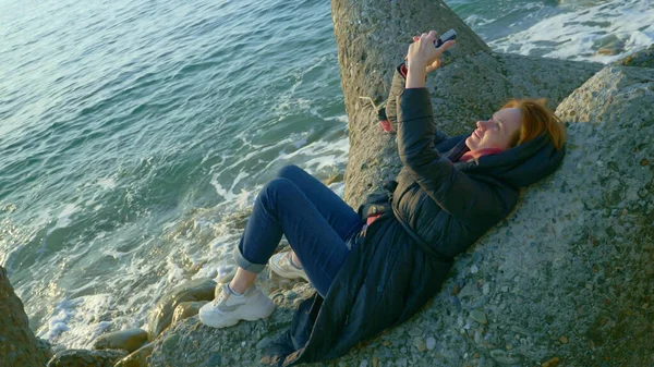 Mujer en un abrigo utiliza un teléfono inteligente mientras está sentado en las rocas junto al mar frío — Foto de Stock