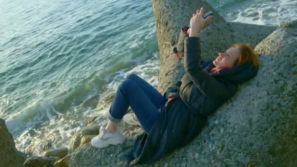 Woman in a coat uses a smartphone while sitting on the rocks by the cold sea — Stock Video