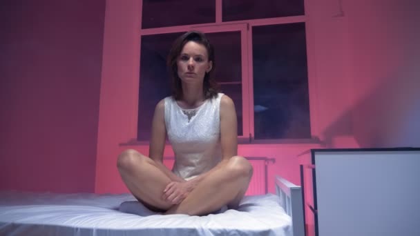 Eine taube Frau in einem weißen Kleid sitzt auf einem Bett in einem verrauchten Raum — Stockvideo