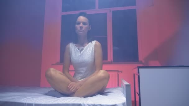 Оніміла жінка в білій сукні сидить на ліжку в димчастій кімнаті — стокове відео