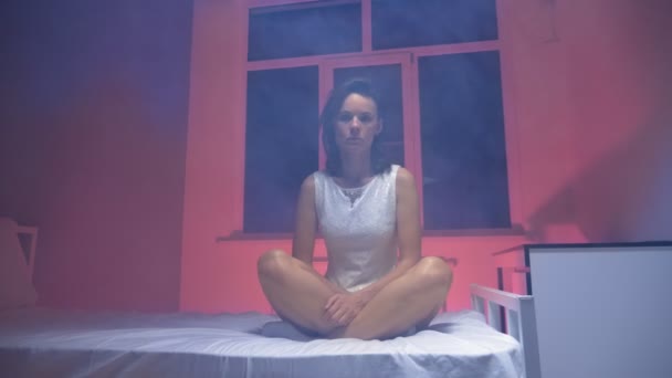 Оніміла жінка в білій сукні сидить на ліжку в димчастій кімнаті — стокове відео