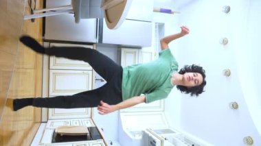 dikey video. Yakışıklı, kıvırcık adam mutfakta neşeyle dans ediyor. 