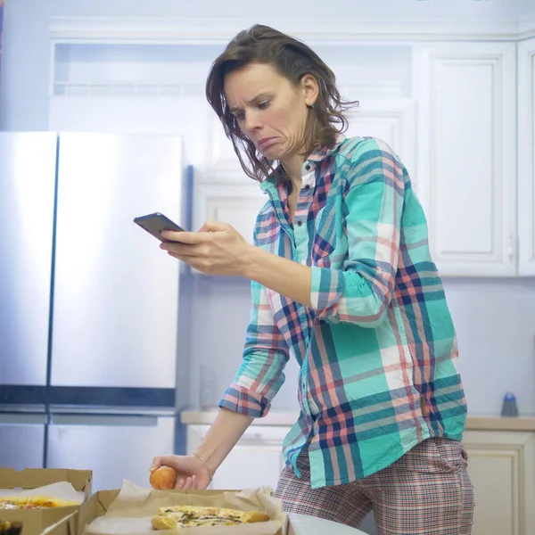 Chica usando el teléfono inteligente mientras se sienta en casa en su cocina — Foto de Stock