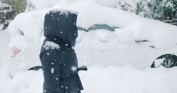 En kvinna med pensel för att rengöra snö från en bil står framför en snötäckt bil under ett snöfall — Stockvideo