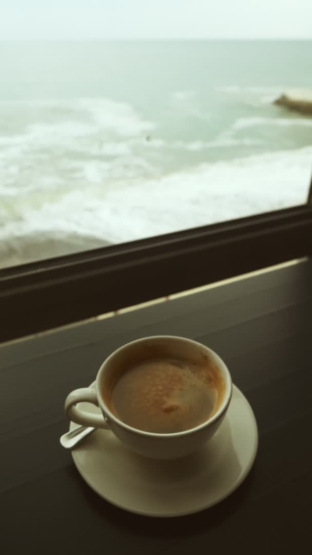Vertikální video. detail hrnečku s kávou na stole na pozadí okna s výhledem na bouřlivé moře — Stock video