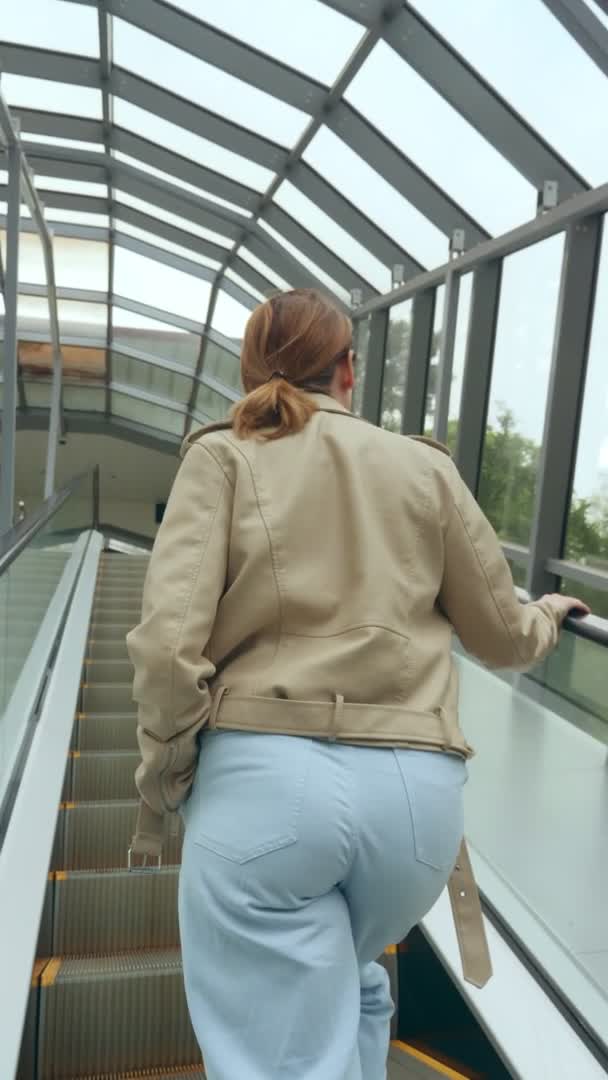 Vídeo vertical. visão traseira, mulher de jaqueta de couro e jeans azuis em uma escada rolante em um túnel transparente — Vídeo de Stock
